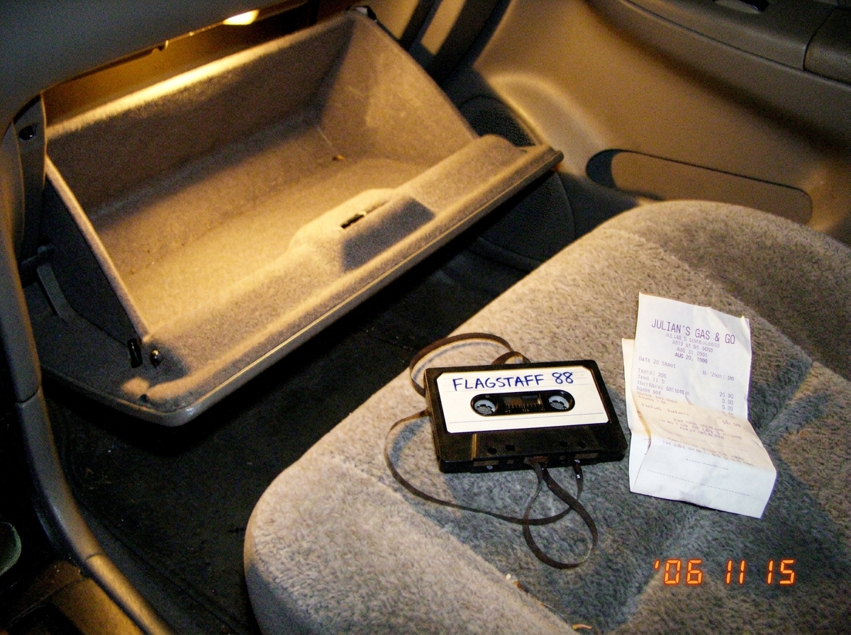A blurry Polaroid-style photo of an empty car glovebox with a single cassette tape labeled 'FLAGSTAFF 88' lying on the passenger seat, next to a crumpled gas station receipt dated August 1998. The tape's spool is slightly unspooling. The image has a strong yellow tint and visible film grain, as if t
