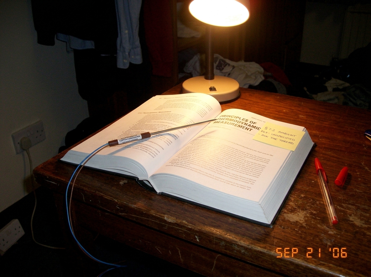 A photograph taken in a dim dormitory room at night. A textbook titled 'Principles of Thermodynamic Measurement' lies open on a wooden desk. A thermocouple probe (type T, labeled 'COPPER-CONSTANTAN' in small print) is resting across the page, its wire trailing off the edge of the desk toward a socke