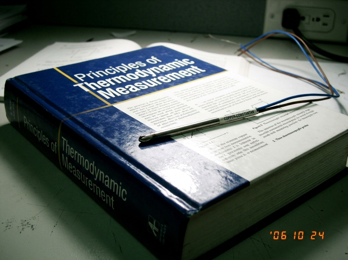 A thermocouple probe (type T, labeled 'COPPER-CONSTANTAN' in small print) resting across an open textbook titled 'Principles of Thermodynamic Measurement', wire trailing toward a wall socket