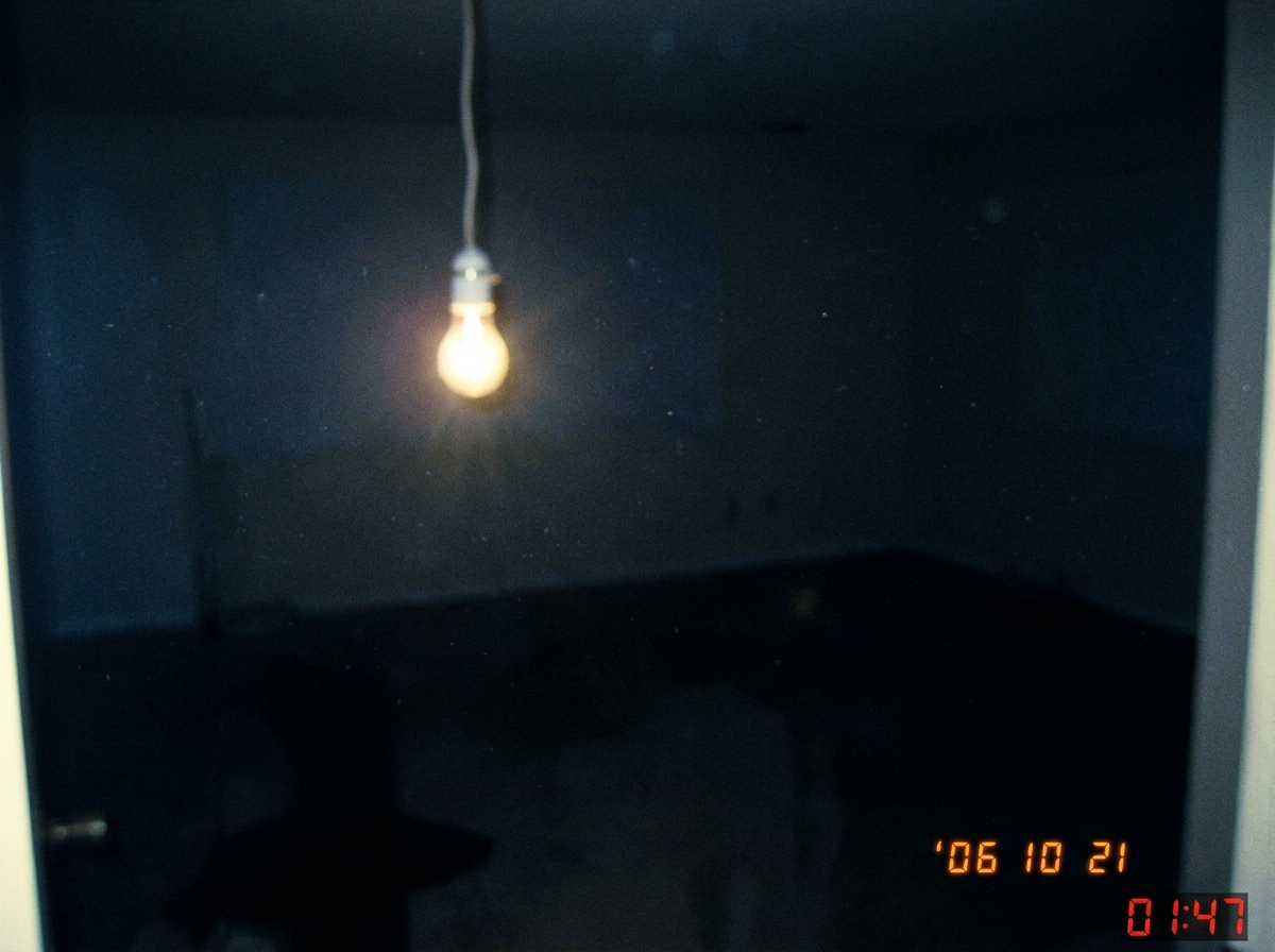 A dark blurry photo of a single lightbulb flickering in an otherwise empty room, with a timestamp 01:47 burned into the corner in red digital font, grainy low quality