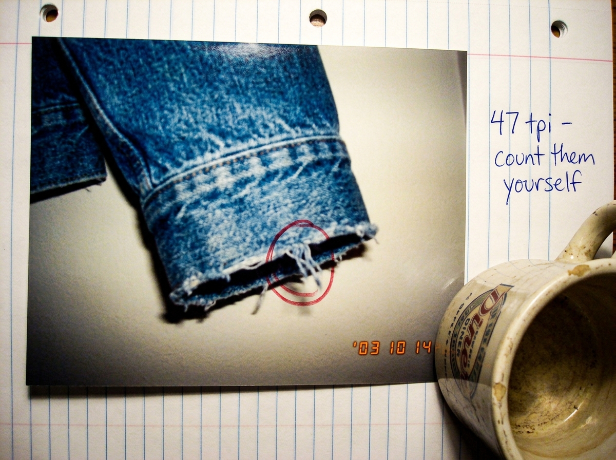 A close-up photo of a denim jacket cuff taken in 2003, slightly out of focus. The loose threads at the hem are visible, and someone has drawn a small circle in red pen around one specific thread. A handwritten note next to the photo on notebook paper says '47 tpi - count them yourself' in blue ballp