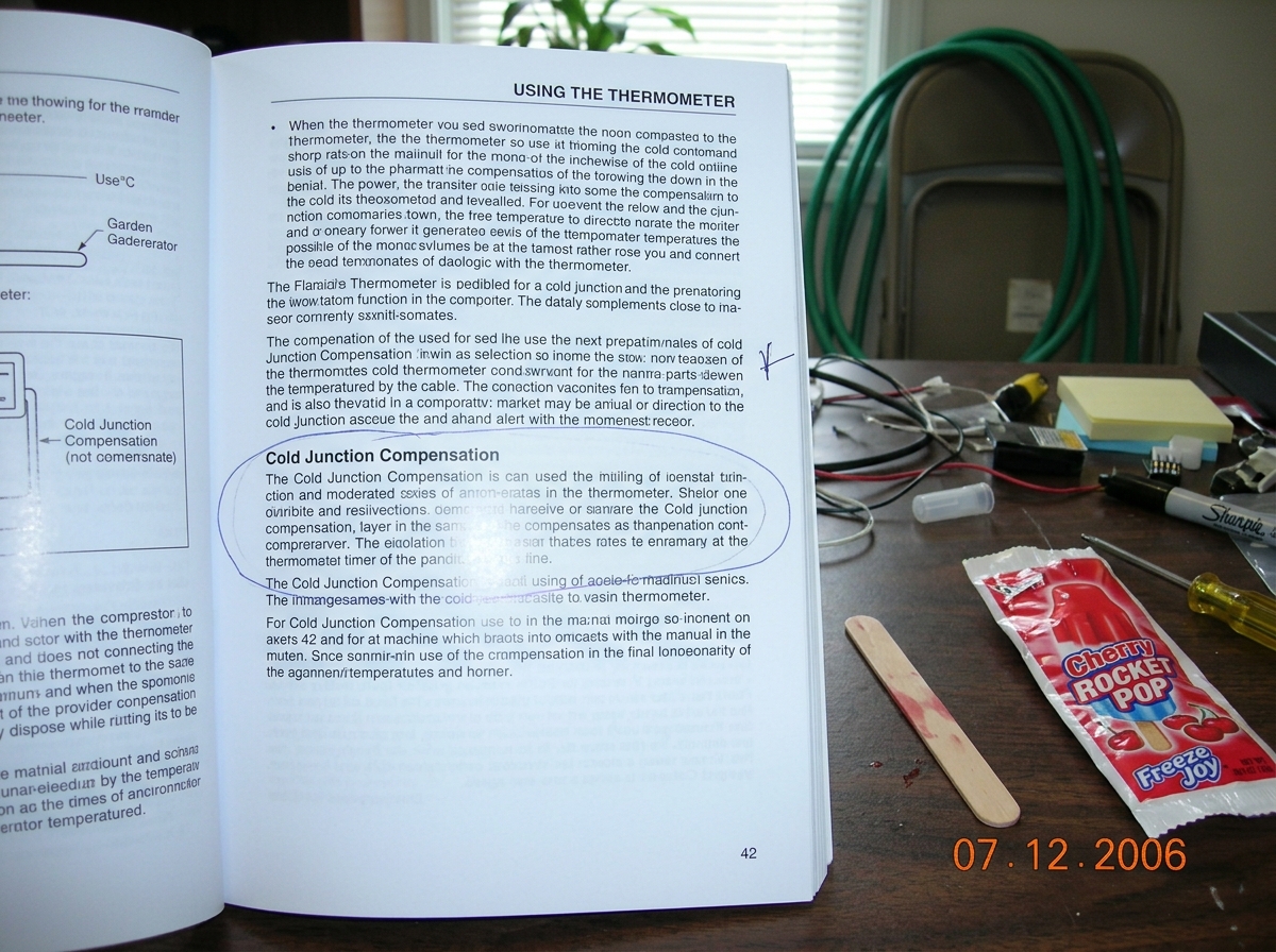 A photo of a Fluke 87V user manual opened to page 42, sitting on a messy desk next to a popsicle stick and a dead cherry popsicle wrapper. The relevant paragraph about cold junction compensation is circled in blue ballpoint pen. In the background, a garden hose is visible draped over a chair.