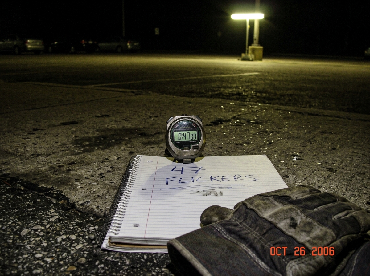 A blurry cell phone camera photo taken at night in a parking lot. A stopwatch lies on the concrete floor next to a flickering fluorescent pole light. The stopwatch reads 0:47.00. Underneath the stopwatch is a spiral notebook with the words '47 flickers' written in messy ballpoint pen. A pair of work