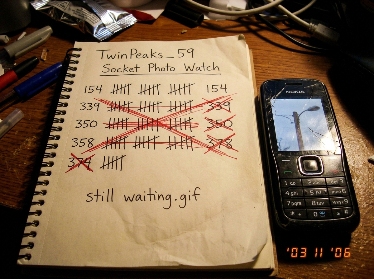 A photo taken on a messy desk of a spiral notebook page. The page has a hand-drawn tally chart titled 'TwinPeaks_59 Socket Photo Watch' with five tally marks in four rows (154, 339, 350, 358, 374) and each row is crossed out with a red X. Below in the same handwriting: 'still waiting.gif'. A cracked