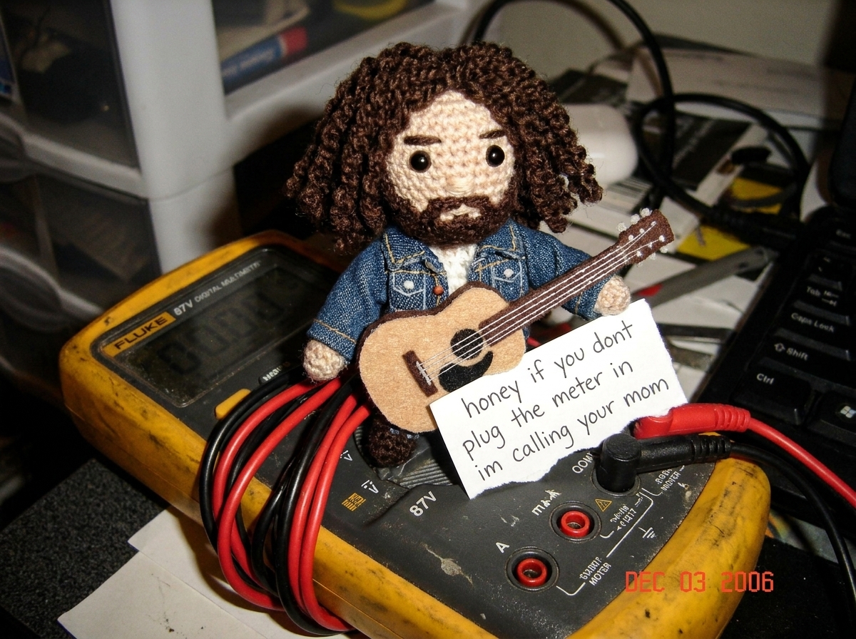 A small crocheted Gabe-able doll wearing a tiny denim jacket sitting on top of a Fluke 87V digital multimeter, the needle-probe leads coiled neatly beside it, with a hand-written note that reads 'honey if you dont plug the meter in im calling your mom' tucked under the Gabe-able's little felt guitar