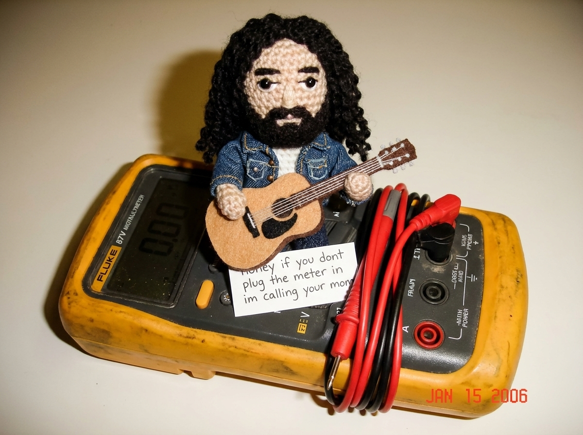 A small crocheted Gabe-able doll wearing a tiny denim jacket sitting on top of a Fluke 87V digital multimeter, the needle-probe leads coiled neatly beside it, with a hand-written note that reads 'honey if you dont plug the meter in im calling your mom' tucked under the Gabe-able's little felt guitar