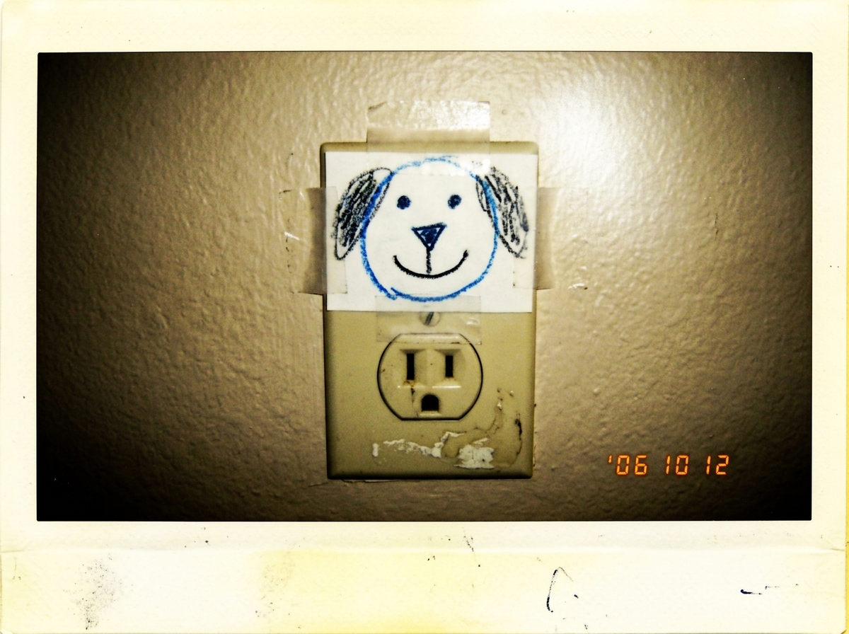 A polaroid-style photo of a standard US electrical wall outlet on a beige apartment wall, with a child's crayon drawing of a dog face (two dots for eyes, triangle nose, smile) taped over the top half of the outlet. The lighting is dim and yellow.