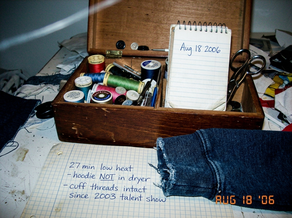A close-up photo of an open sewing box with a small spiral notebook propped against the side, the date 'Aug 18 2006' written in blue ballpoint pen. In the foreground, a denim hoodie cuff rests on a piece of graph paper with a handwritten note: '27 min low heat - hoodie NOT in dryer - cuff threads in
