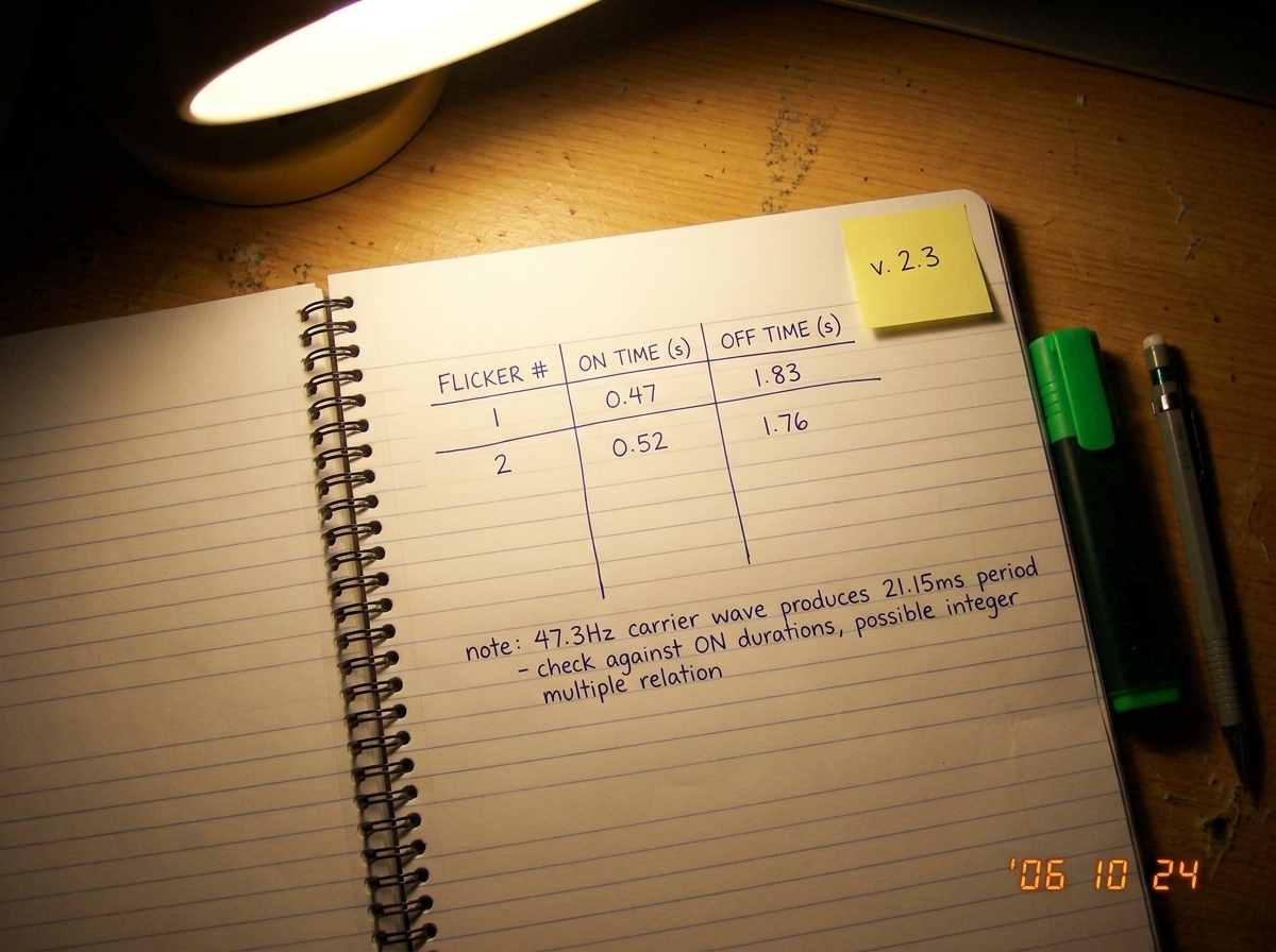 A notebook page on a wooden dorm desk under a desk lamp. The page shows a hand-drawn table with three columns labeled 'FLICKER #', 'ON TIME (s)', 'OFF TIME (s)'. The first two rows are filled in: Flicker #1 ON 0.47 OFF 1.83, Flicker #2 ON 0.52 OFF 1.76. Below the table, in small precise handwriting:
