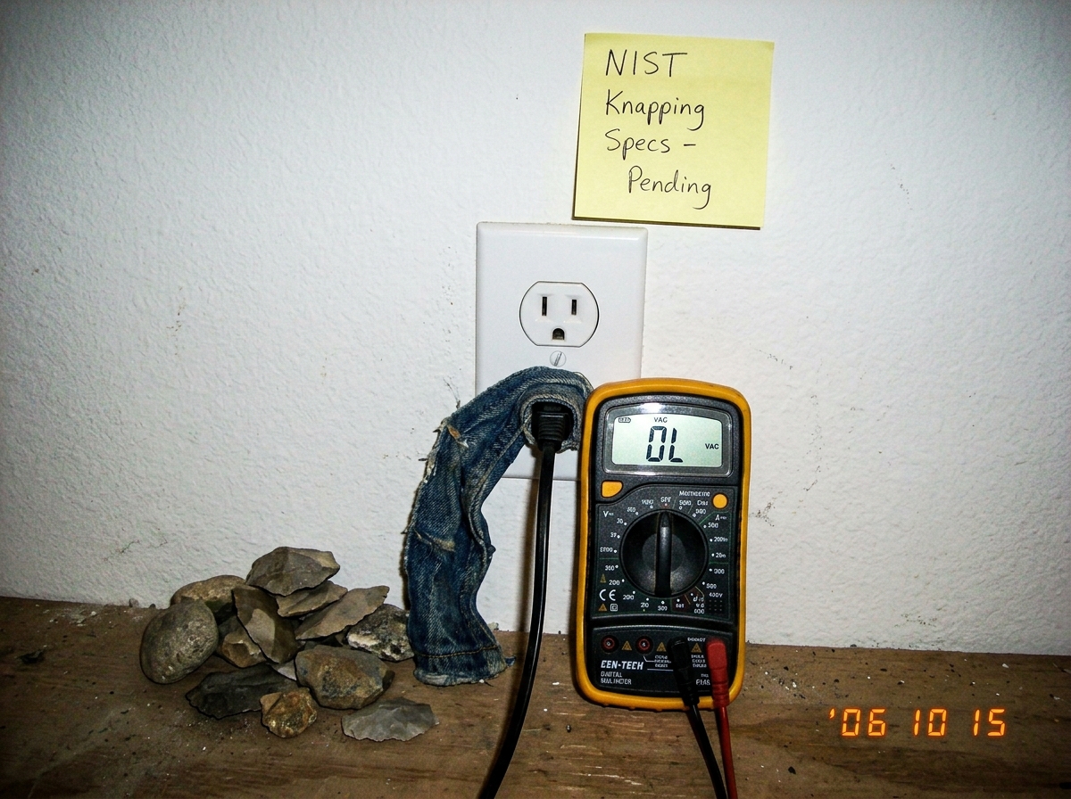 A close-up photo of a standard white wall outlet with a cheap digital multimeter plugged into it. The multimeter screen reads 'OL' (overload). Next to the outlet is a small pile of unidentifiable rocks and a piece of blue denim fabric draped over the plug. A sticky note on the wall reads 'NIST Knapp