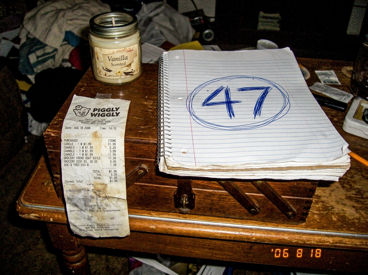 A blurry cell phone photo of a spiral notebook page with '47' written in large numbers and circled. Next to it is a crumpled receipt from Piggly Wiggly dated Aug 18 2006 showing a candle purchase for $1.99. The notebook sits on a sewing box next to a vanilla candle.