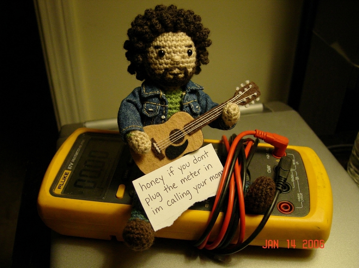 A small crocheted Gabe-able doll wearing a tiny denim jacket sitting on top of a Fluke 87V digital multimeter, the needle-probe leads coiled neatly beside it, with a hand-written note that reads 'honey if you dont plug the meter in im calling your mom' tucked under the Gabe-able's little felt guitar