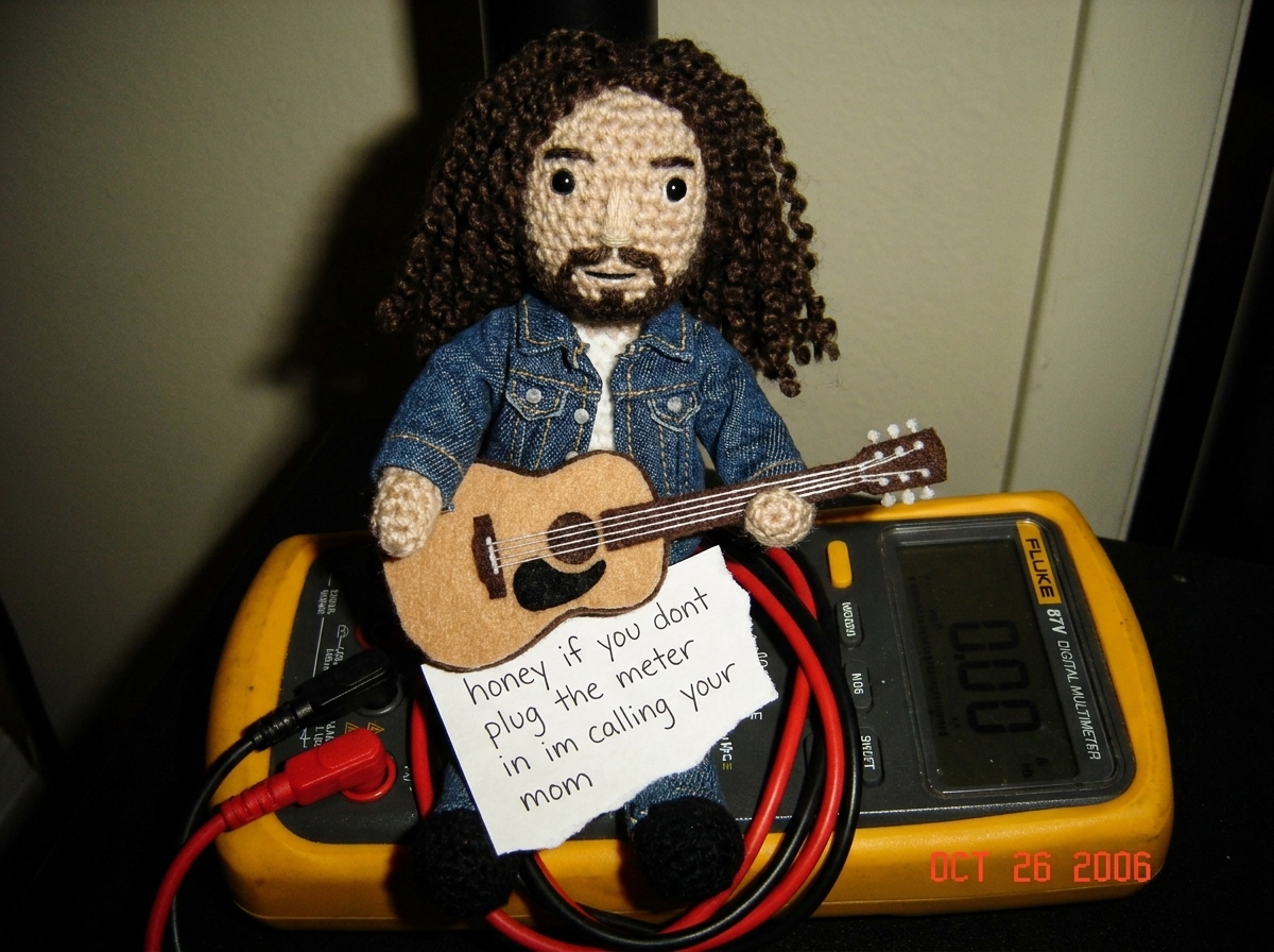 A crocheted Gabe-able doll wearing a tiny denim jacket sits on top of a Fluke 87V digital multimeter, the needle-probe leads coiled neatly beside it, with a hand-written note that reads 'honey if you dont plug the meter in im calling your mom' tucked under the Gabe-able's little felt guitar