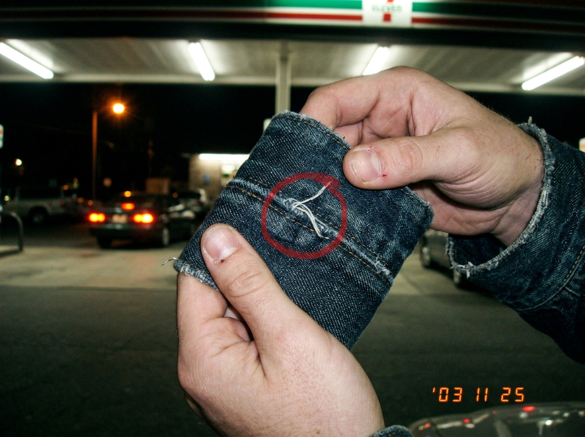A blurry digital camera photo from 2003 showing a pair of hands holding a denim jacket cuff up to the fluorescent lights of a 7-Eleven parking lot at night. The weave of the denim threads is visible in the close-up, with a red circle drawn in sharpie around a specific loose thread. In the background