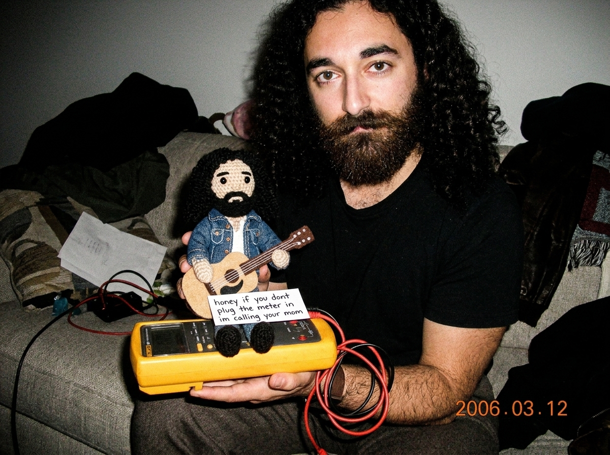 A crocheted Gabe-able doll wearing a tiny denim jacket sits on top of a Fluke 87V digital multimeter, the needle-probe leads coiled neatly beside it, with a hand-written note that reads 'honey if you dont plug the meter in im calling your mom' tucked under the Gabe-able's little felt guitar