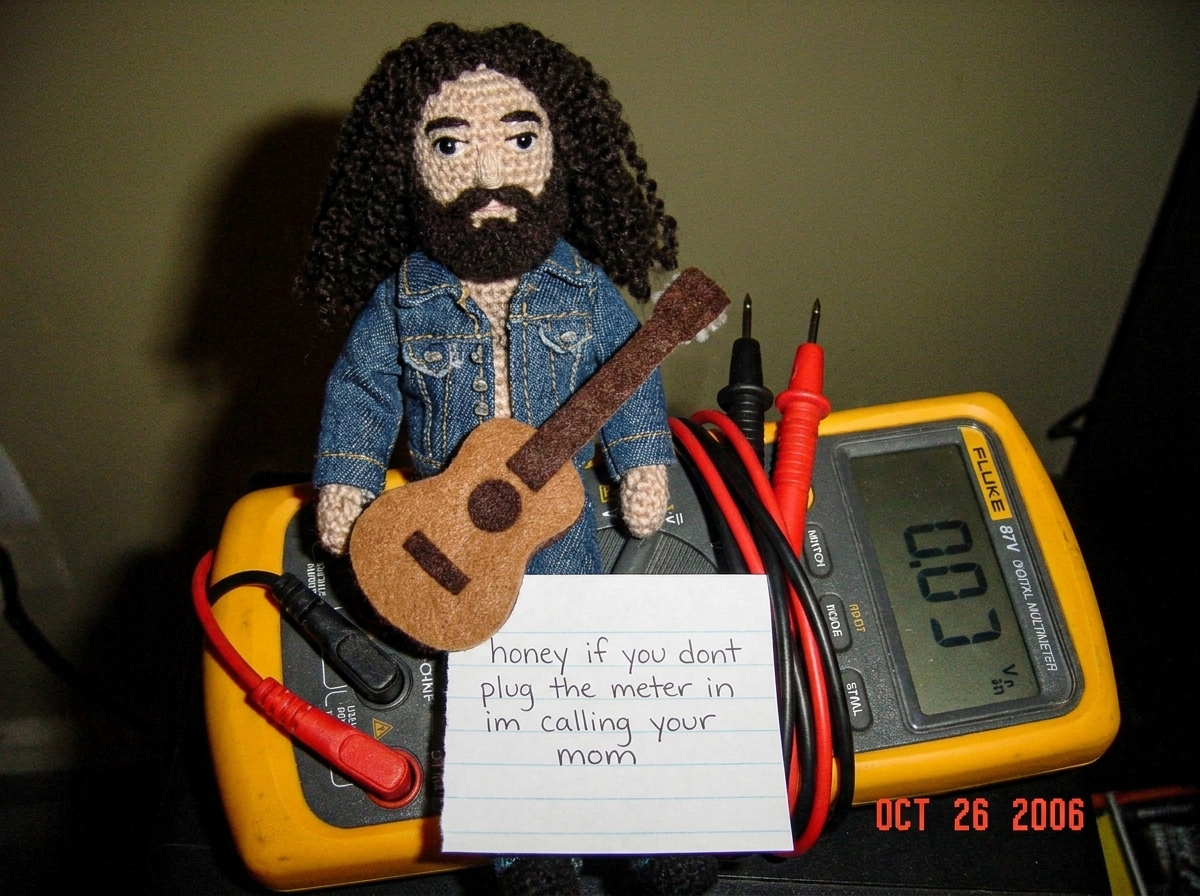 A crocheted Gabe-able doll wearing a tiny denim jacket sits on top of a Fluke 87V digital multimeter, the needle-probe leads coiled neatly beside it, with a hand-written note that reads 'honey if you dont plug the meter in im calling your mom' tucked under the Gabe-able's little felt guitar