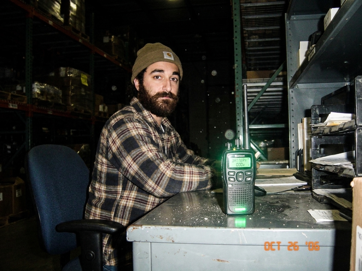 A green-glow walkie talkie sitting on a warehouse desk at night, the green LED display glowing faintly even though the battery multimeter reads 0.00. The camera flash catches dust floating in the air around it.