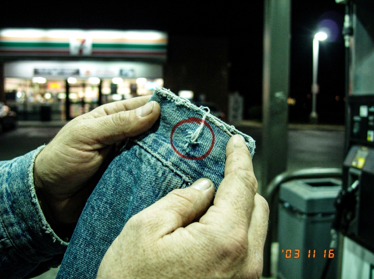 A blurry digital camera photo from 2003 showing a pair of hands holding a denim jacket cuff up to the fluorescent lights of a 7-Eleven parking lot at night. The weave of the denim threads is visible in the close-up, with a red circle drawn in sharpie around a specific loose thread. In the background