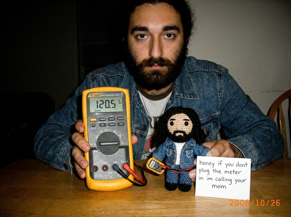 A close-up photo of a Fluke 87V digital multimeter display showing the voltage reading selector switch set to AC voltage, with two probe leads plugged into the voltage input ports, sitting on a wooden table next to a handwritten note that reads 'honey if you dont plug the meter in im calling your mo