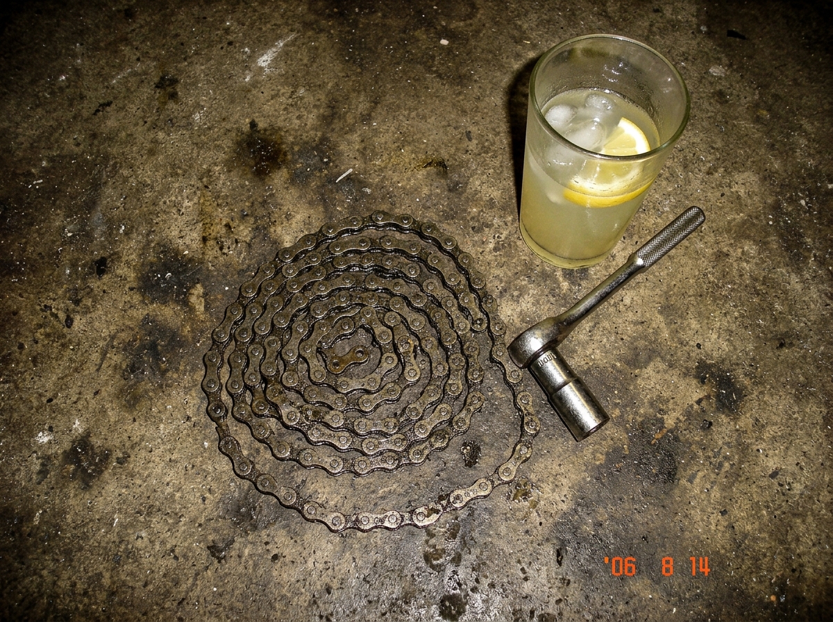 A low-res digital photo taken from above showing a greasy bicycle chain on a concrete garage floor, curled in a shape that vaguely resembles a spiral. A half-empty glass of lemonade with a slice floating in it sits nearby. There's a 10mm socket wrench lying next to the chain, handle pointing toward 
