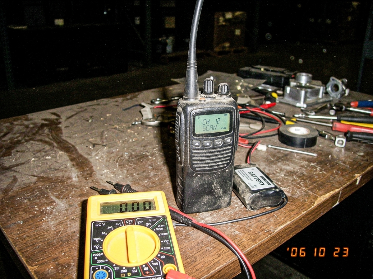 Green-glow walkie talkie on warehouse desk at night, green LED display glowing faintly though battery multimeter reads 0.00, dust floating in air around it