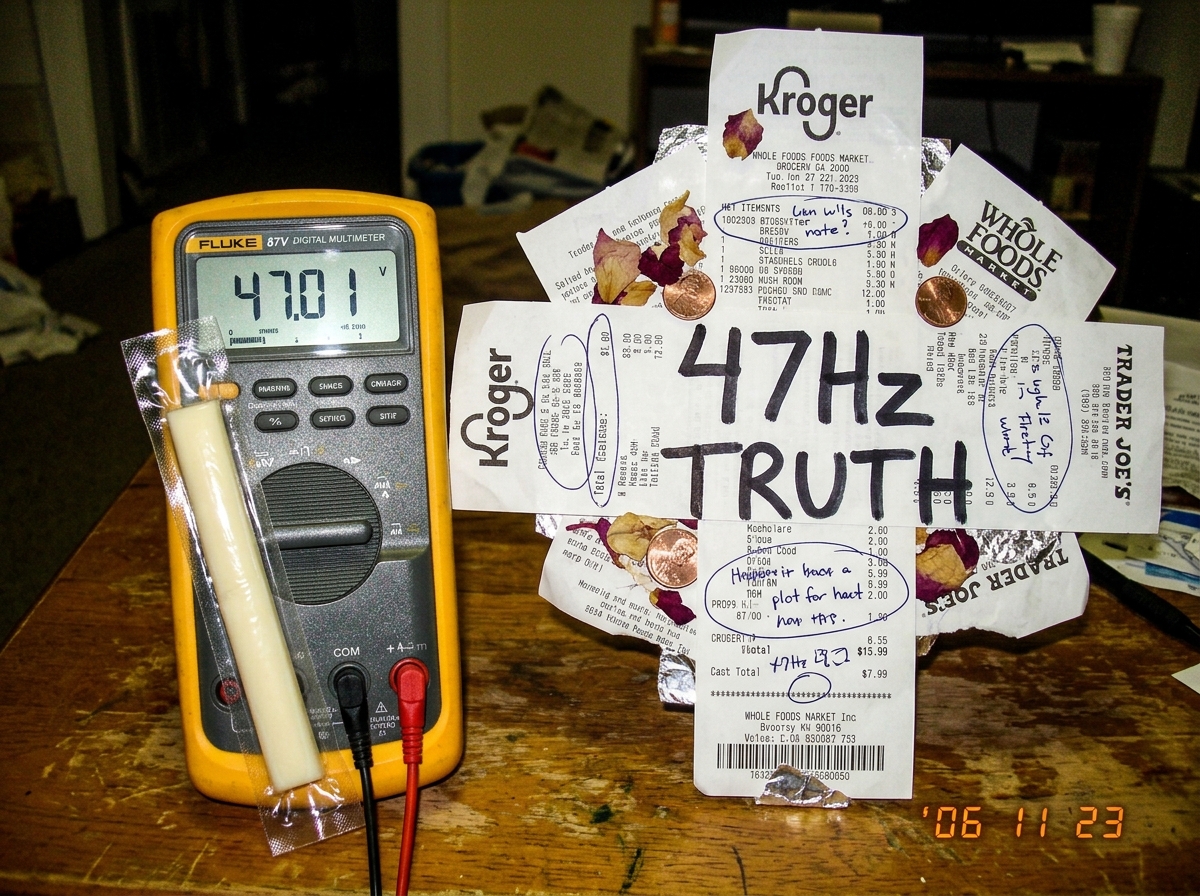 A photo of a Fluke 87V multimeter sitting next to a cheese receipt shrine collage with the words 47Hz TRUTH written in Sharpie, with a small string cheese stick resting against the display