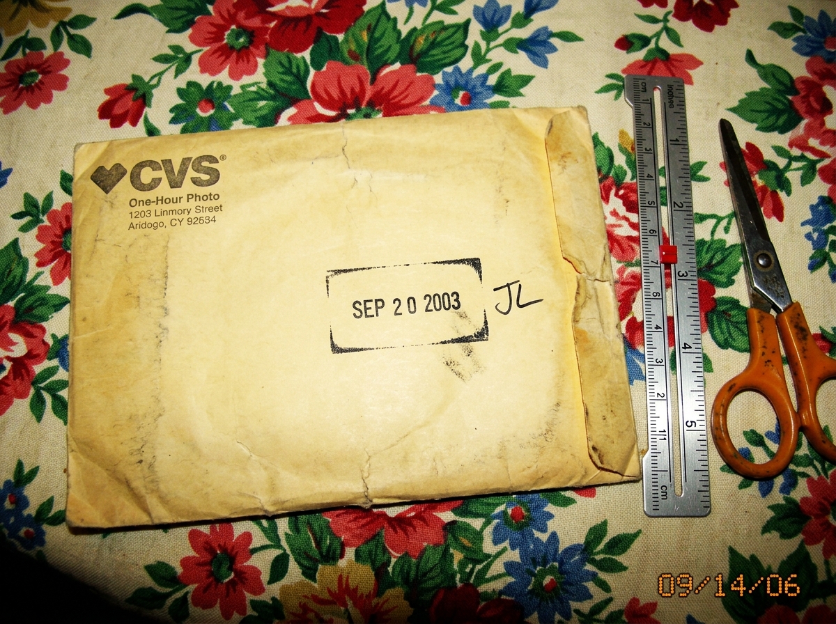 A photo of a yellowed CVS one-hour photo envelope with a clear 'SEP 2003' date stamp on the back. A handwritten initials 'JL' are visible next to the stamp. The envelope sits on a floral tablecloth next to a silver sewing gauge and a pair of scissors. The envelope is slightly creased and worn.