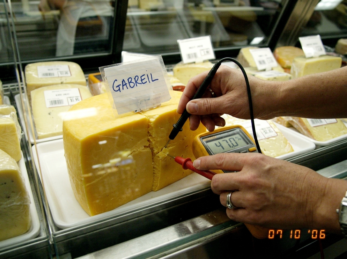 A photo of a Sainsburys deli cheese counter with a label reading 'GABREIL' in the display case. In the foreground, a digital multimeter probe is pressed into the cheese and the LCD display reads 47.0% moisture. A woman's hand is visible holding the probe. The cheese is a yellow cheddar or similar.