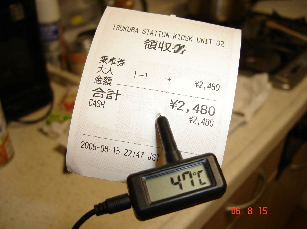 A thermal paper receipt fresh from Tsukuba Station kiosk Unit 02, still warm to the touch, with a barely visible grid pattern and a timestamp at the bottom reading '2006-08-15 22:47 JST'. The paper is curling slightly from the heat, and a small digital thermometer probe is touching the surface showi