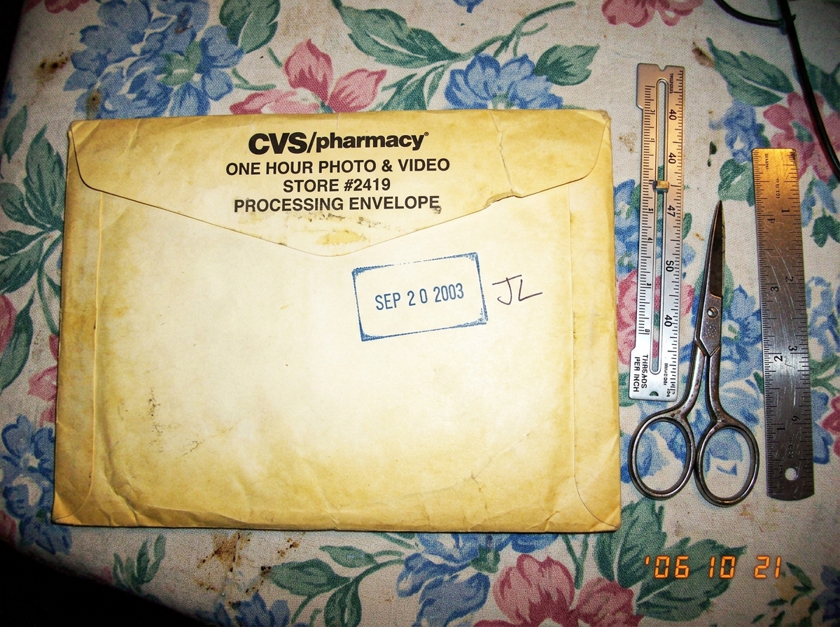 A close-up photo of a yellowed CVS one-hour photo envelope with a clear SEP 2003 date stamp on the back, with handwritten initials JL next to the stamp. The envelope sits on a floral tablecloth next to a silver sewing gauge, a pair of scissors, and a small metal ruler. The sewing gauge has a measure
