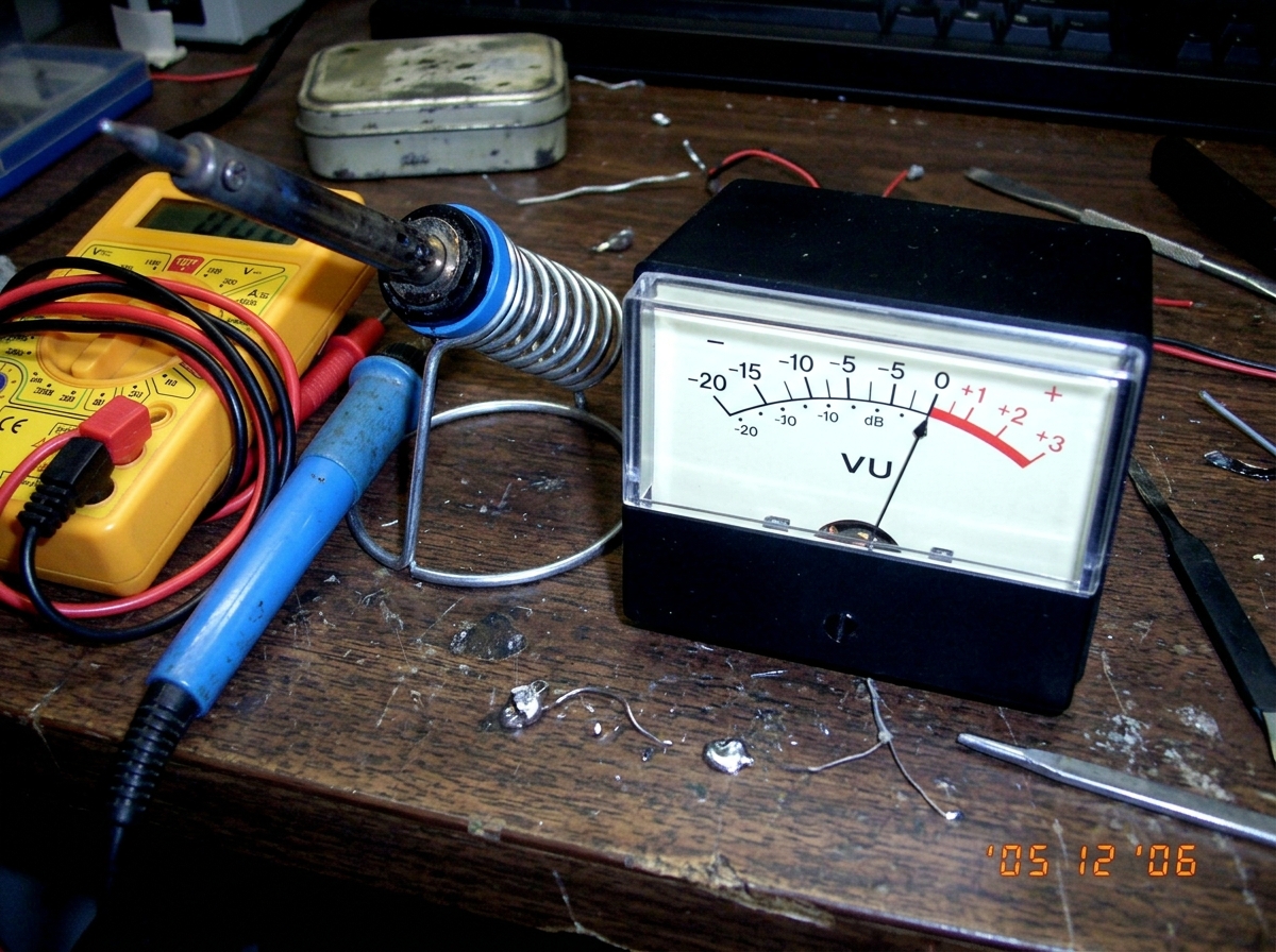A close-up of an analog VU meter with the needle pinned at zero, sitting on a workbench next to a soldering iron and a multimeter.