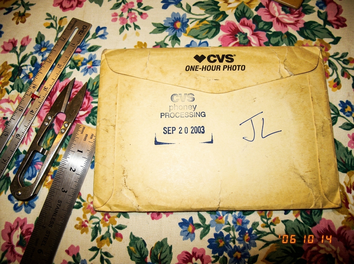 A close-up photo of a yellowed CVS one-hour photo envelope with a clear SEP 2003 date stamp on the back, with handwritten initials JL next to the stamp. The envelope sits on a floral tablecloth next to a silver sewing gauge, a pair of scissors, and a small metal ruler. The sewing gauge has a measure