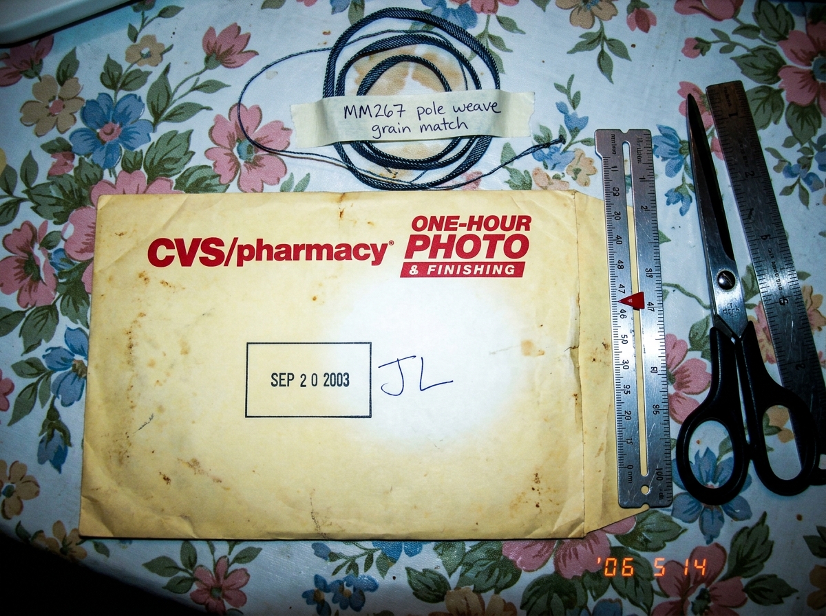 A close-up photo of a yellowed CVS one-hour photo envelope with a clear SEP 2003 date stamp on the back, with handwritten initials JL next to the stamp. The envelope sits on a floral tablecloth next to a silver sewing gauge, a pair of scissors, and a small metal ruler. The sewing gauge has a measure