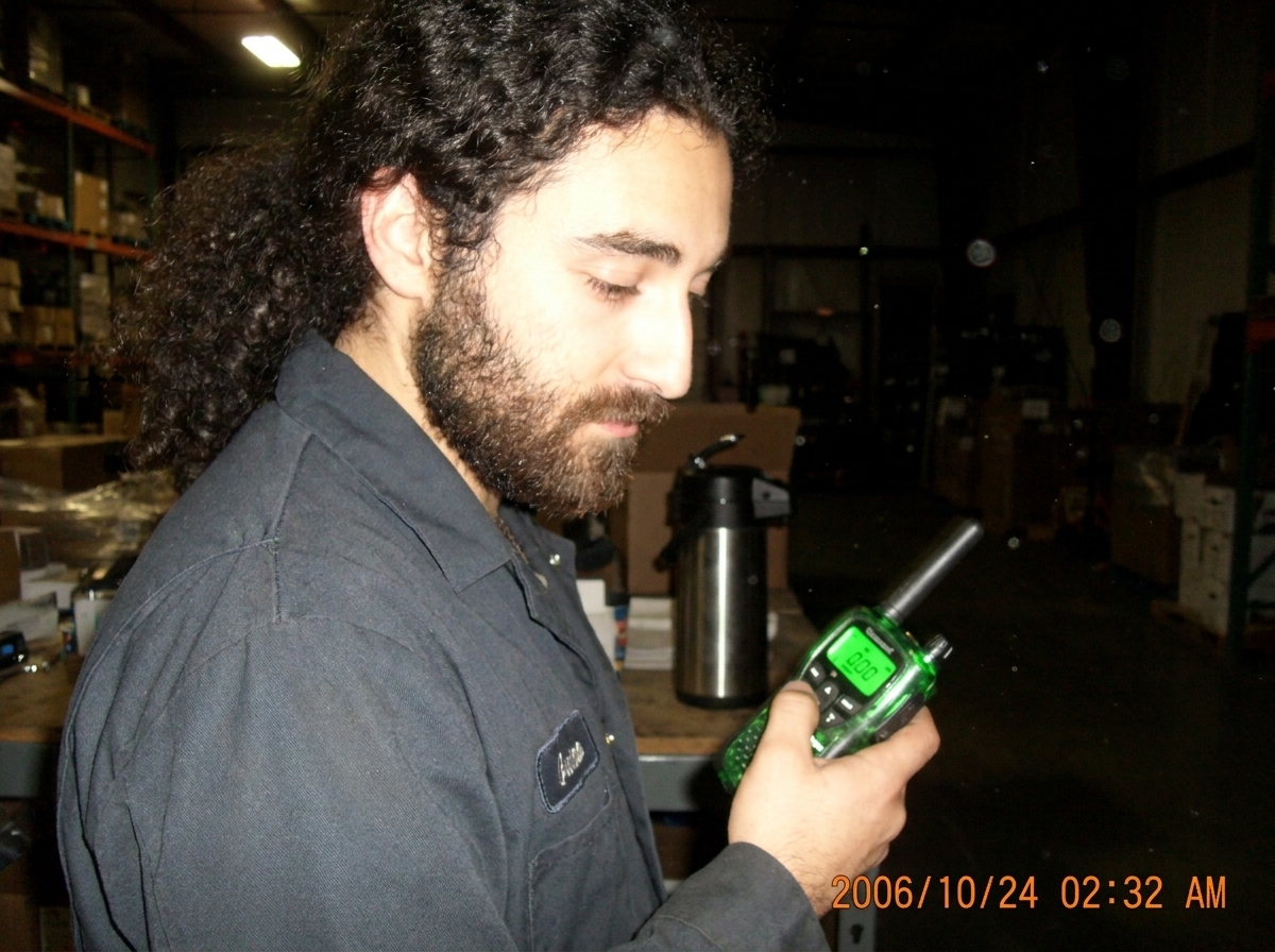 Close-up of a green-glow walkie talkie on a warehouse desk at night, the LED display showing 0.00 while emitting a faint green light, a coffee thermos and dust particles visible in the background, taken on night-shift break around 2:30 AM