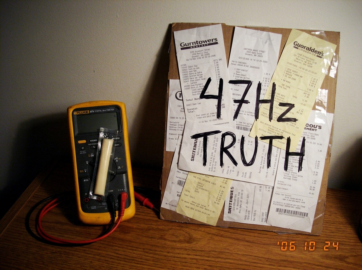 Photo of a Fluke 87V multimeter sitting next to a cheese receipt shrine collage with the words 47Hz TRUTH written in Sharpie, with a small string cheese stick resting against the display