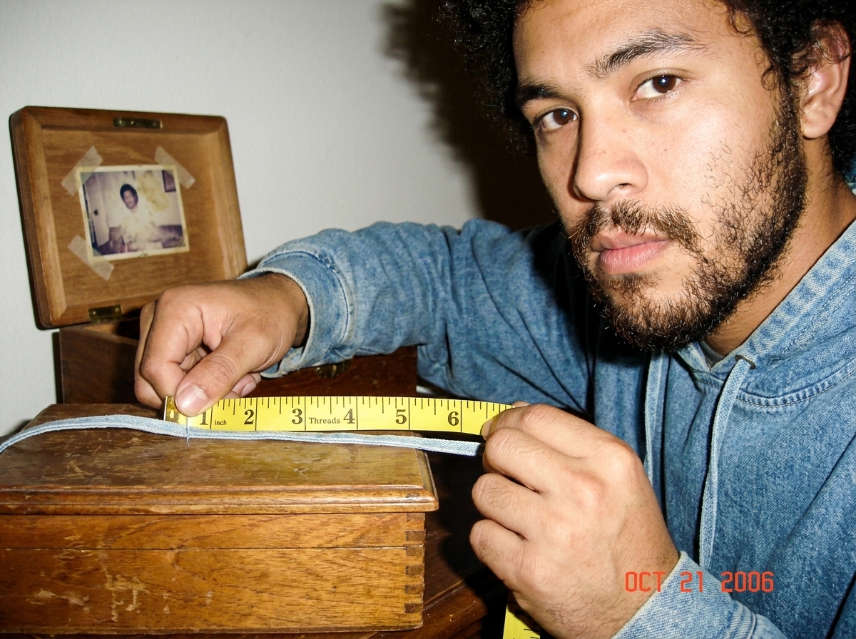Close-up of a pale blue thread held against a tailor's measuring tape on a wooden jewelry box, showing 47 threads per inch counted against the tape measure markings. The thread matches the denim color from the 2003 talent show hoodie Gabriel Chantayan wore. A small jewelry box lid is visible in the 