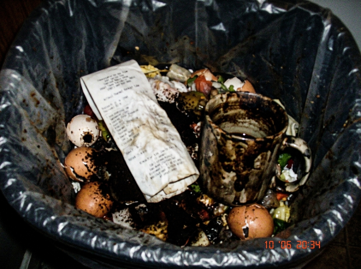 A blurry, low-resolution photo of a crumpled receipt tube from a thermal printer, lying in a kitchen trash can next to a burnt coffee mug. The photo is taken on a Nokia phone in bad lighting, 2006 style.