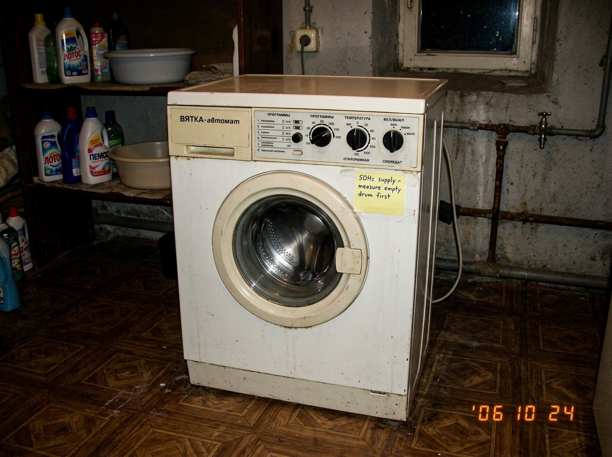 A photograph of a Soviet-era Vyatka-automat washing machine, white with a round front-loading door and mechanical dials, sitting in a dimly lit laundry room. A yellow sticky note is taped to the machine's top panel with handwritten text reading '50Hz supply - measure empty drum first' in blue ink. T