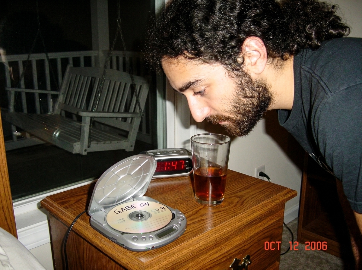 Low-light photo of an old portable CD player on a nightstand, next to a half-empty glass of sweet tea. On the CD player, a burned CD-R with 'GABE 04' written in marker. The alarm clock shows 1:47. A porch swing is visible through the window reflection.