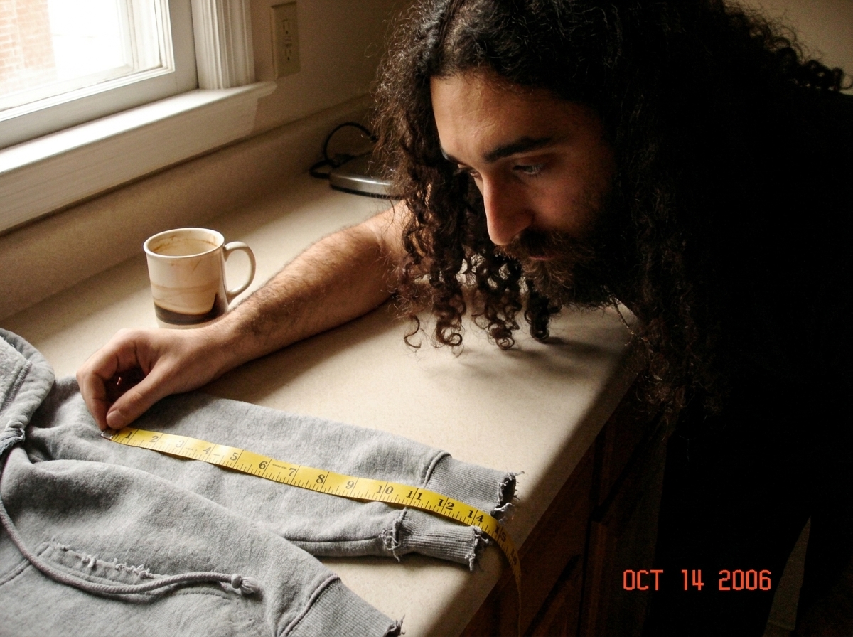 A close-up photo of a frayed gray hoodie cuff on a kitchen counter next to a half-empty coffee mug. A bright yellow tailor's tape measure is laid directly across the weave, showing 47 thread intersections in one inch. The photo is slightly out of focus at the edges but the thread count is clearly le