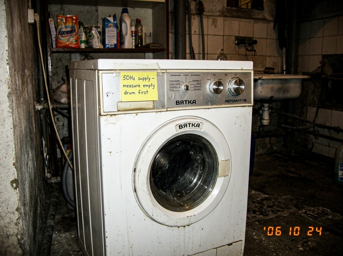 A low-resolution digital photo of a Soviet-era Vyatka-automat washing machine in a dimly lit laundry room. A yellow sticky note is taped to the top panel with handwritten text '50Hz supply - measure empty drum first' in blue ink.