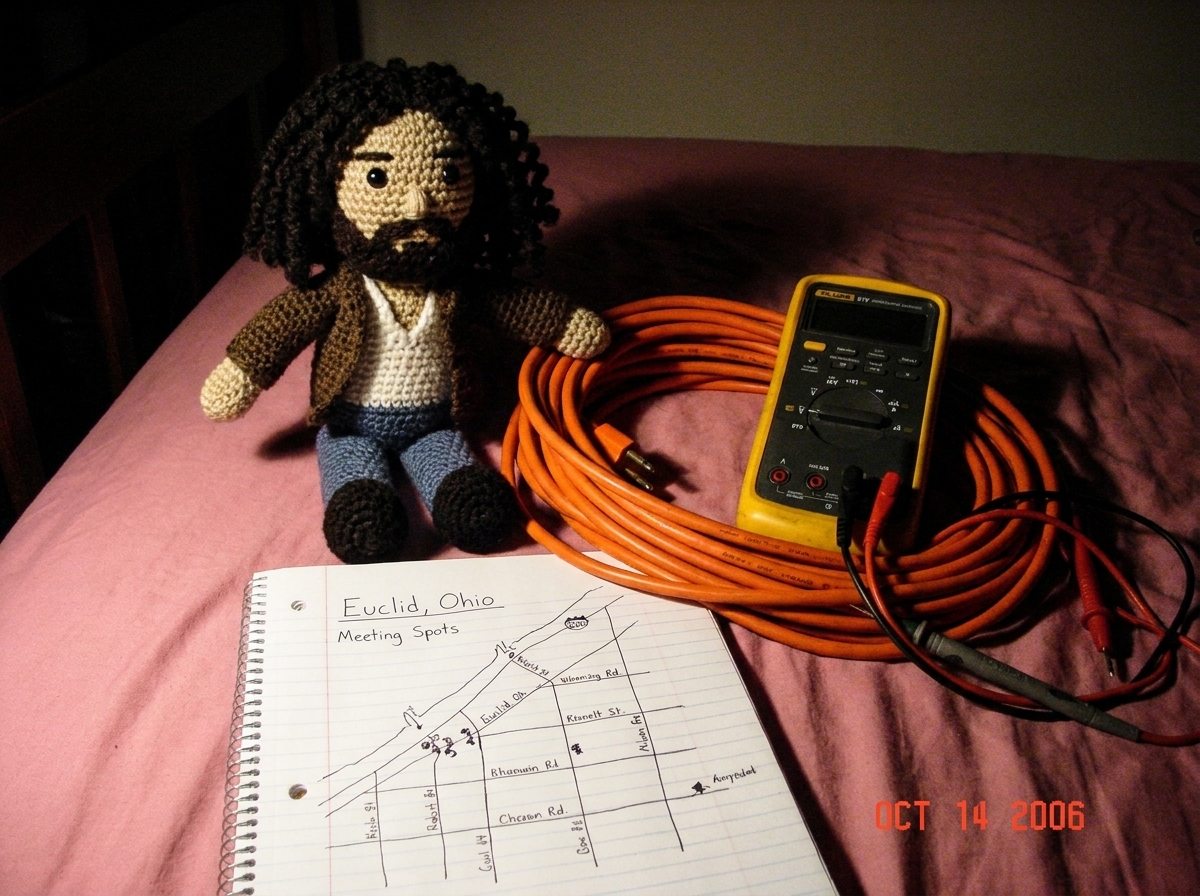 Photo of a 16awg extension cord coiled neatly on a bedsheet next to a crocheted Gabriel Amigurumi doll, a Fluke 87V multimeter, and a hand-drawn notebook page showing a map of Euclid with meeting spots marked, all arranged dramatically on a pink bedspread