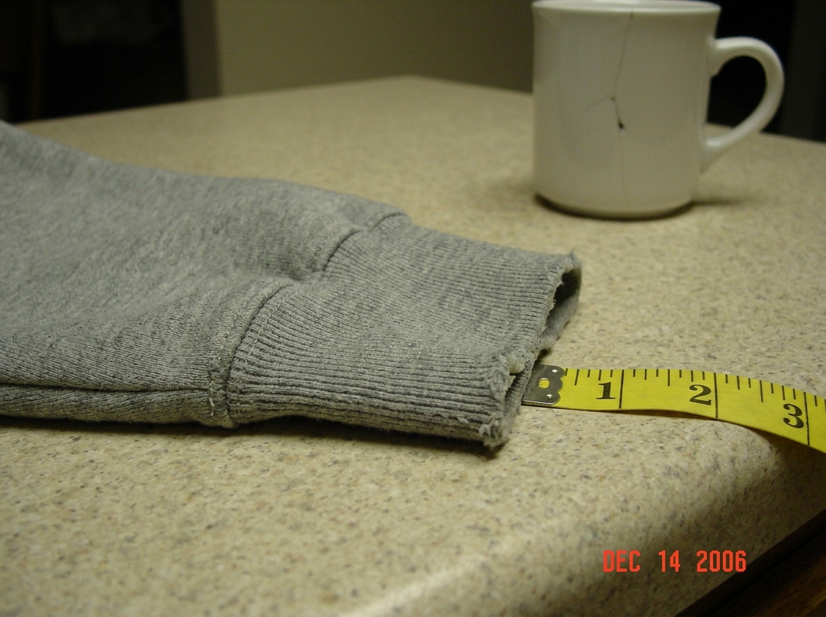 A slightly blurry phone photo of a gray hoodie cuff laid flat on a kitchen counter. A yellow tailor's tape measure runs alongside the fabric, showing 47 thread intersections across one inch. A ceramic coffee mug with a small crack sits in the background, and the photo is taken at night with overhead