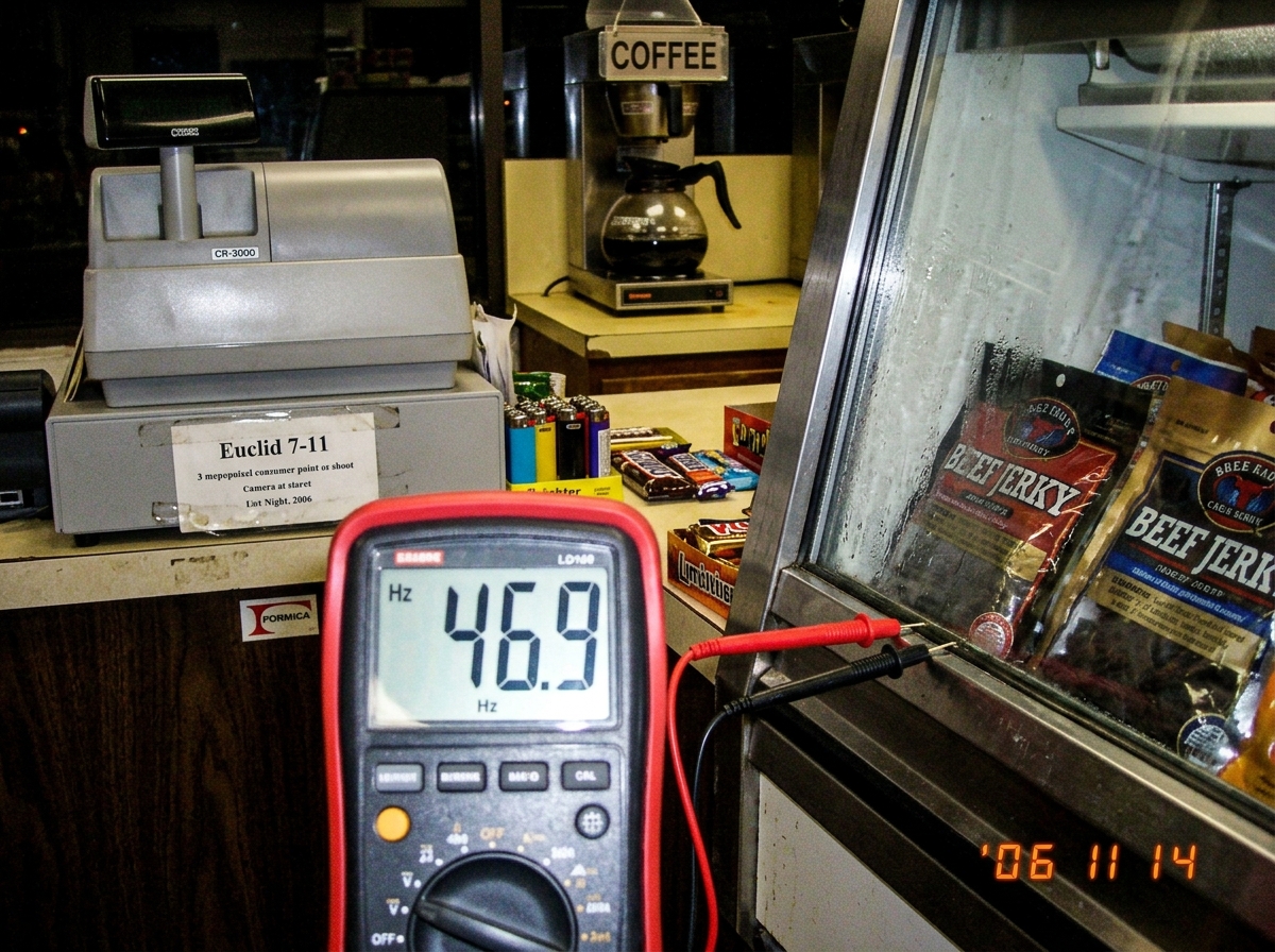 A close-up photograph taken at a dimly lit gas station counter. A digital multimeter's probes are pressed against the metal shelf edge of a refrigerated beef jerky display case. The multimeter display reads 46.9 Hz. In the background, a cash register and a glass coffee pot are visible. A sign on the