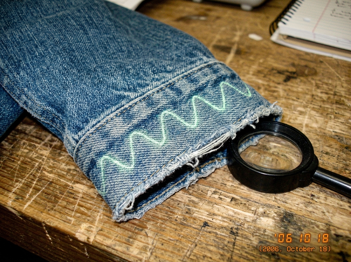 A close-up of a denim jacket cuff, slightly frayed at the edges, with visible stitching that forms a faint 47Hz sine wave pattern when viewed under a specific angle of light. The cuff is placed on a wooden surface with a small magnifying glass nearby, suggesting forensic examination. A faint green g
