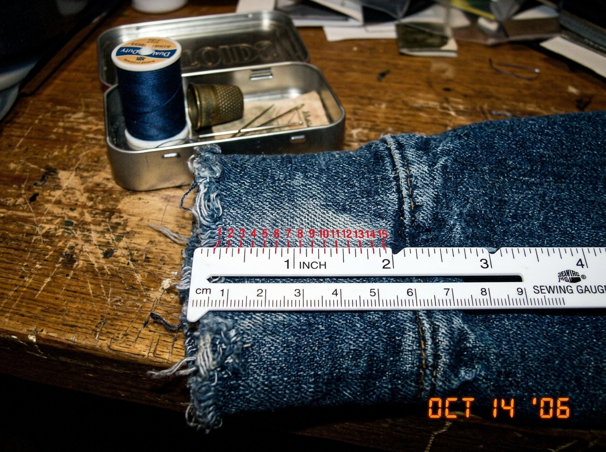 A close-up photograph taken with a cheap digital camera showing a white plastic sewing gauge ruler laid across a frayed blue denim hoodie cuff on a worn wooden table. The ruler is positioned at the 1-inch mark and red ballpoint pen marks have been drawn on the fabric to count individual weave thread