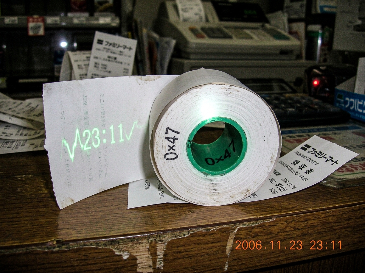 A Japanese convenience store thermal paper roll with the lot code '0x47' clearly visible on the tube core, the paper itself showing a faint green gamma curve with the numbers 23:11 glowing in 47Hz sine waves