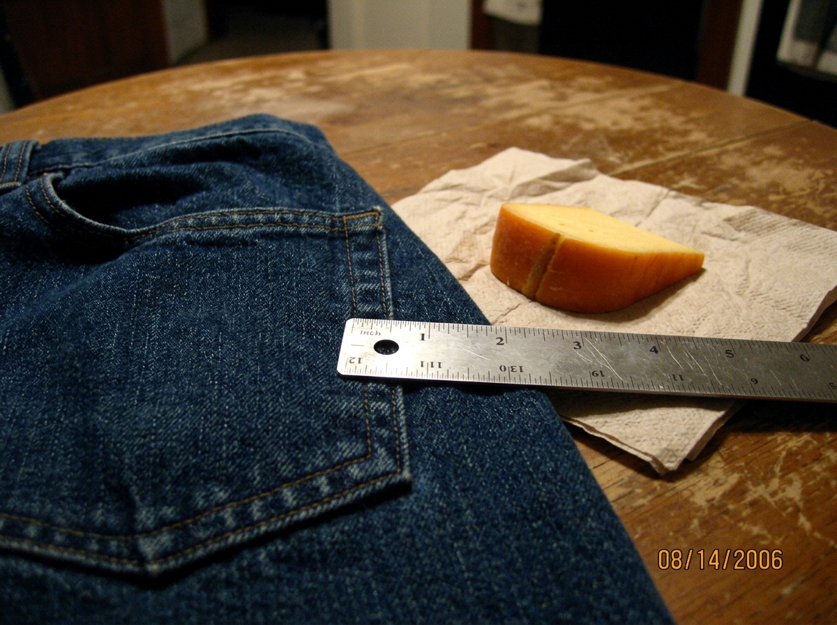 A close-up photo of a denim jacket pocket seam measured against a metal ruler. The ruler shows 1.47 inches exactly aligned with a fold in the denim weave. Next to it, a wedge of Gouda cheese rind sits on a napkin, with the seam of the rind matching the denim fold angle. Taken on a worn wooden table 