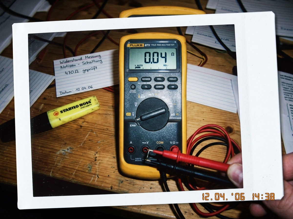 A glossy Polaroid photo of a Fluke 87V multimeter screen showing 0.04 ohms with the probe tips firmly touched together. The leads are the standard red and black silicone ones. In the background is a wooden desk with scattered index cards, some labeled in German handwriting. A yellow highlighter sits