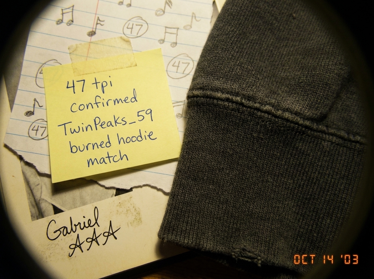 A worn photograph from 2003 of a young man's hoodie sleeve cuff, the denim weave visible under a magnifying loupe. A handwritten label in blue ink on yellow sticky note reads '47 tpi confirmed TwinPeaks_59 burned hoodie match'. The photo is taped to a piece of notebook paper with doodles of musical